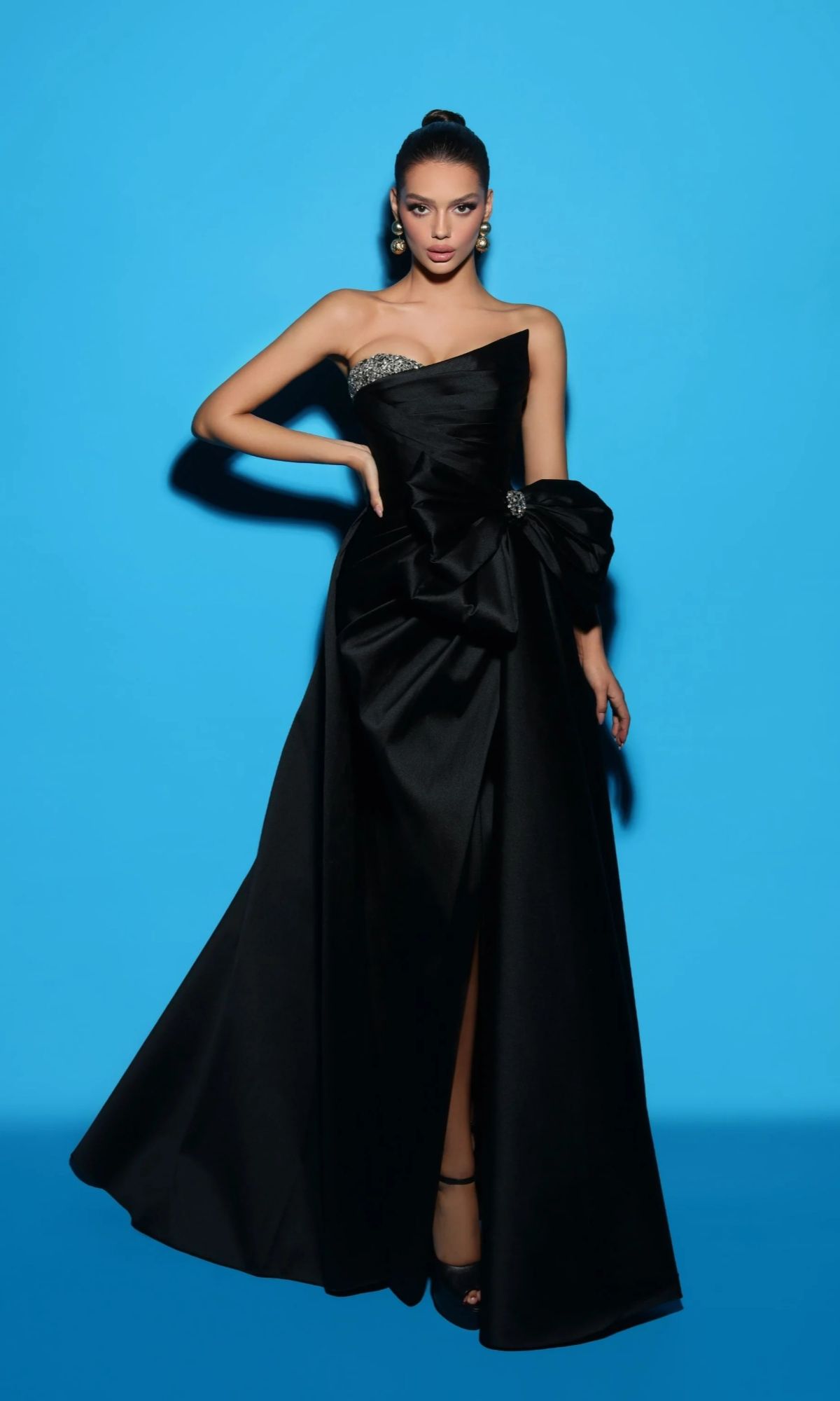 A woman stands against a blue background wearing the Long Formal Dress: Tarik Ediz 98421 in black, featuring a one-shoulder design with a large bow, embellishments, an elegant A-line taffeta skirt, and black high heels.